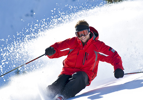 Skier in a red jacket gliding down a snowy slope, leaving a trail of snow spray behind