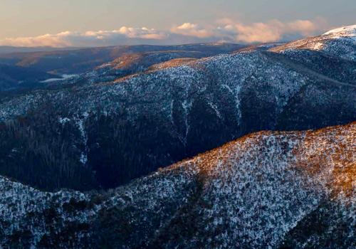 Snow-covered mountain range bathed in the warm glow of a sunset, with layers of forested hills stretching into the distance.