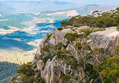 Panoramic view of a rugged cliff overlooking lush green valleys and distant hills, with a serene lake in the background.