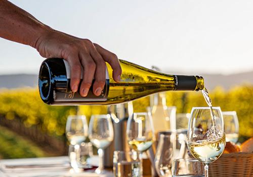 Hand pouring white wine into a glass at sunset, with vineyards and hills in the background.