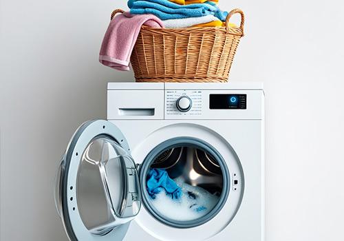 Modern front-loading washing machine with the door open, showing blue laundry inside, and a wicker basket filled with folded clothes on top.