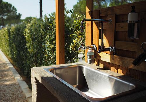 Outdoor sink with a stainless steel basin, dual faucets, and soap dispenser, surrounded by wooden paneling and greenery.