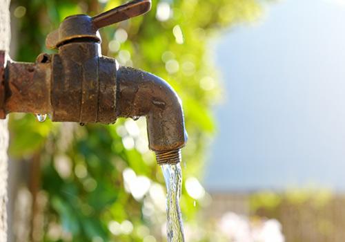 Rustic outdoor faucet with water flowing steadily, set against a backdrop of green foliage and bright sunlight.
